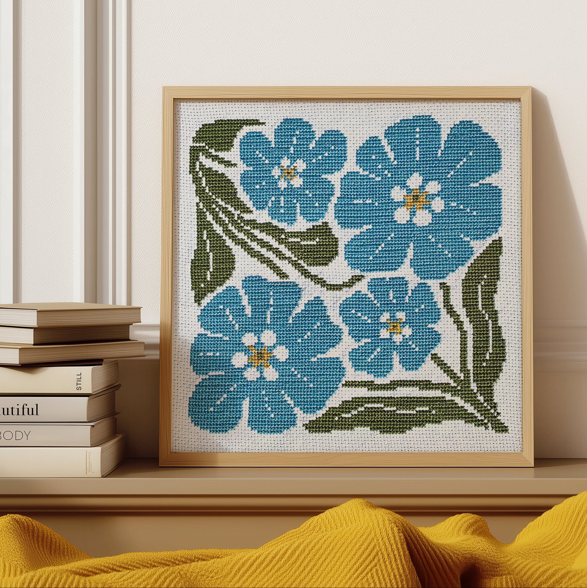 A framed floral needlepoint or cross-stitch artwork with four blue flowers and green leaves on a white canvas, displayed on a wooden shelf next to stacked books against a white paneled wall.