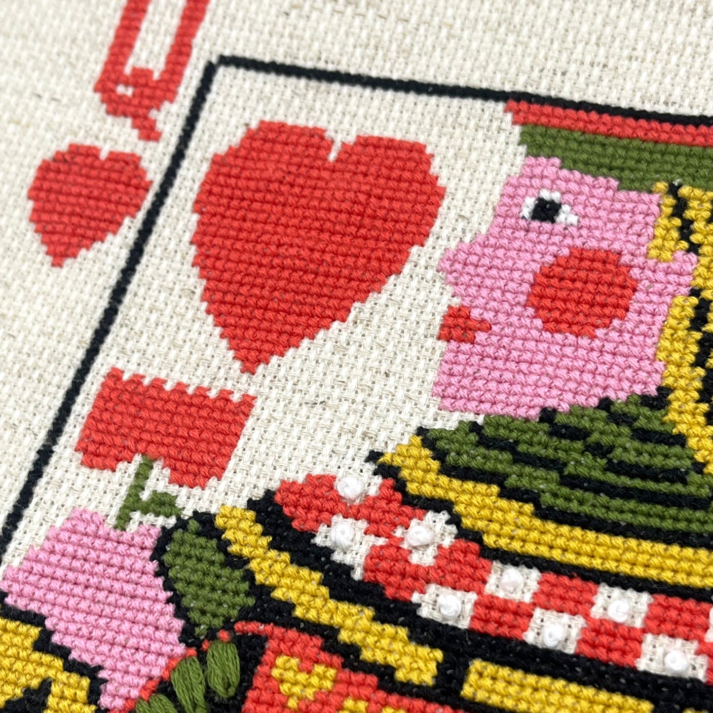 Close-up of a cross-stitch artwork depicting the Queen of Hearts, showing embroidered red hearts, pink face, green hat, and colorful patterned clothing on a cream fabric background.