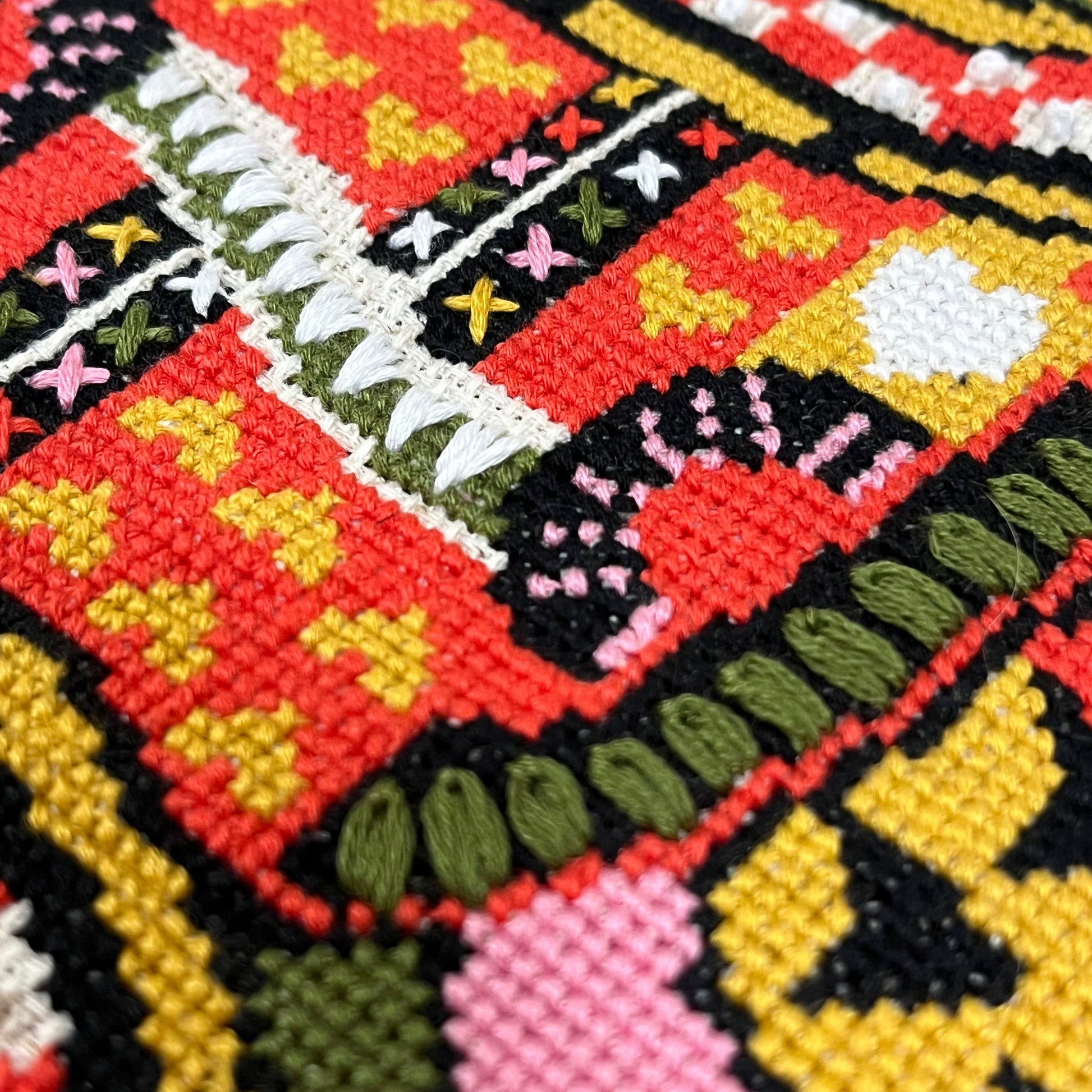 Close-up of a colorful embroidered textile with geometric and floral patterns in red, yellow, pink, green, black, and white.