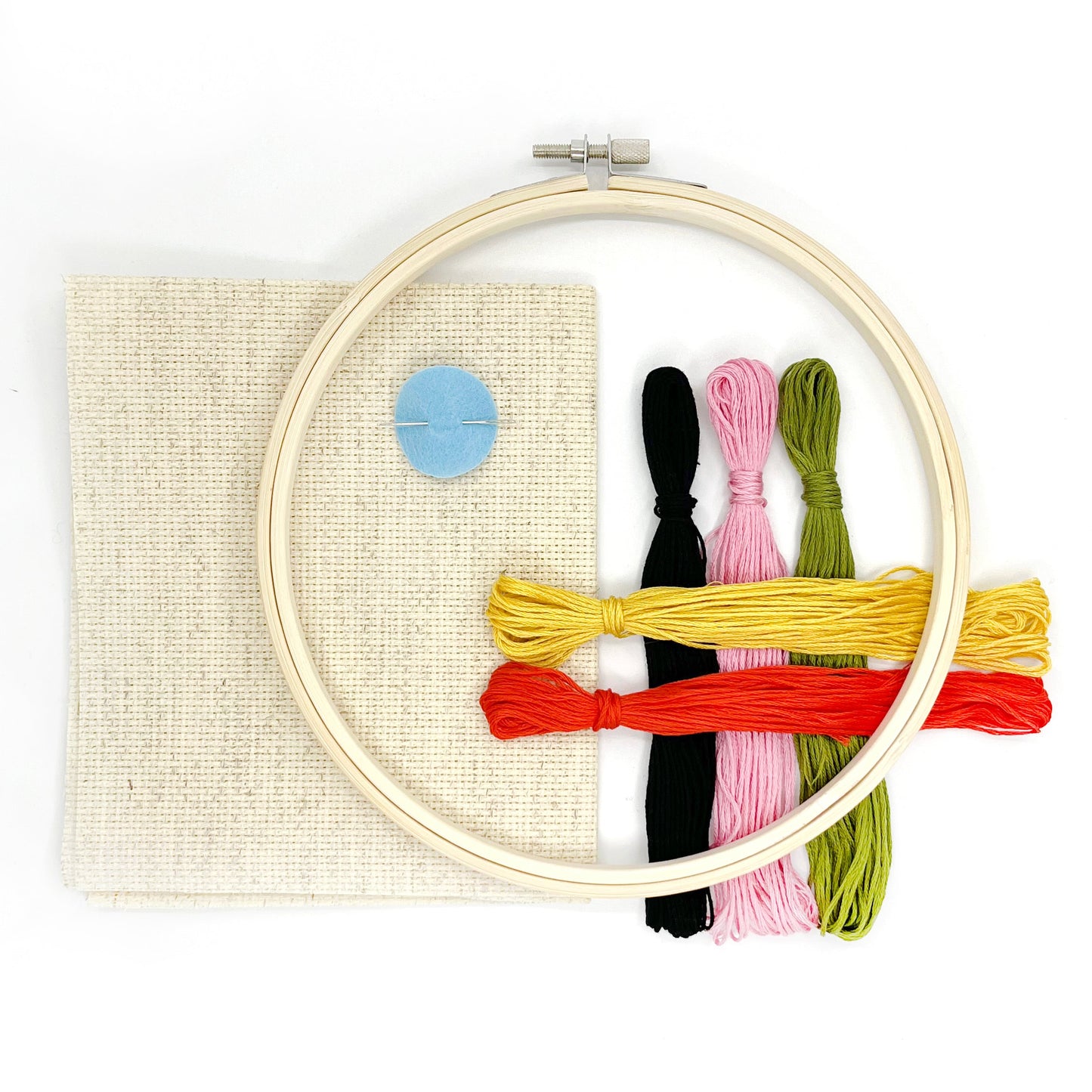 A flat-lay view of a embroidery hoop kit on a white background, containing a wooden embroidery hoop, a beige fabric panel, a blue circular patch, and four colored embroidery threads in black, pink, yellow, and green.
