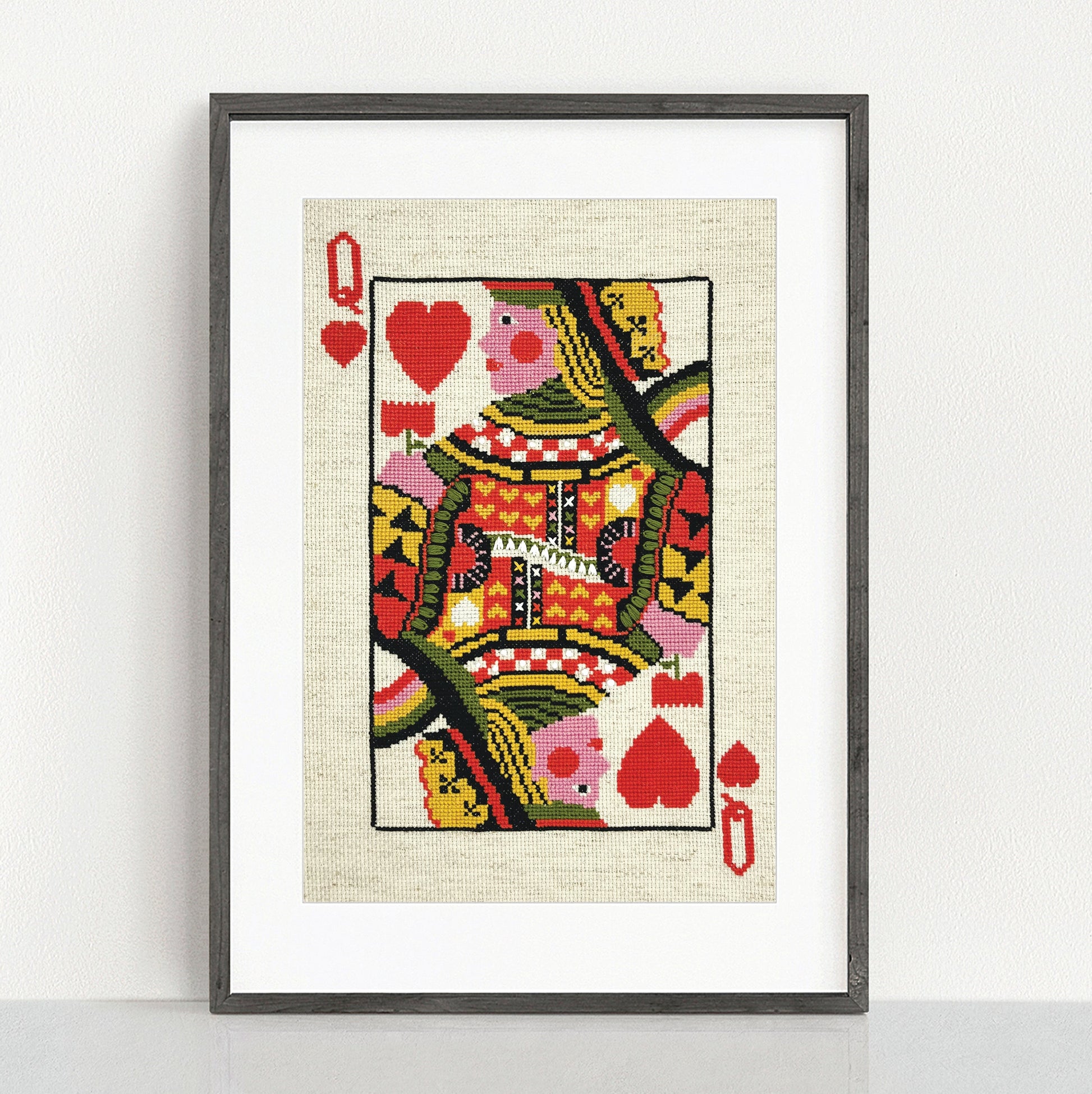 A framed art print of a Queen of Hearts playing card, featuring a vibrant, patterned illustration on a textured cream background, housed in a dark gray frame and leaning against a white wall.