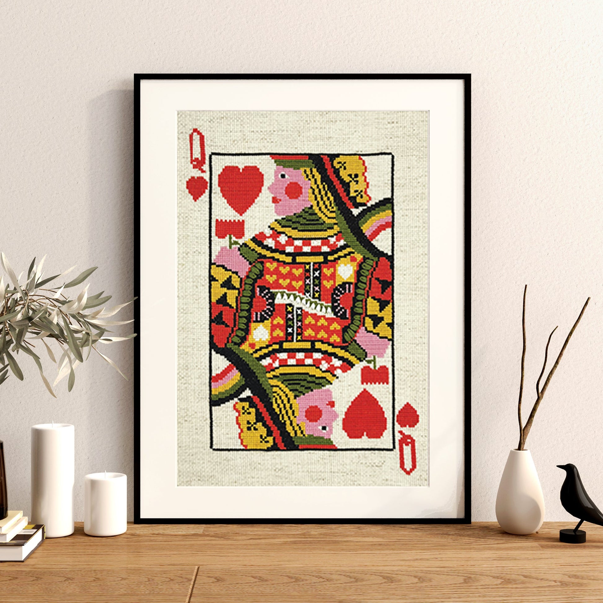 A framed art print of a Queen of Hearts playing card, styled on a wooden shelf next to decorative vases, books, and a small bird figurine against a neutral wall.
